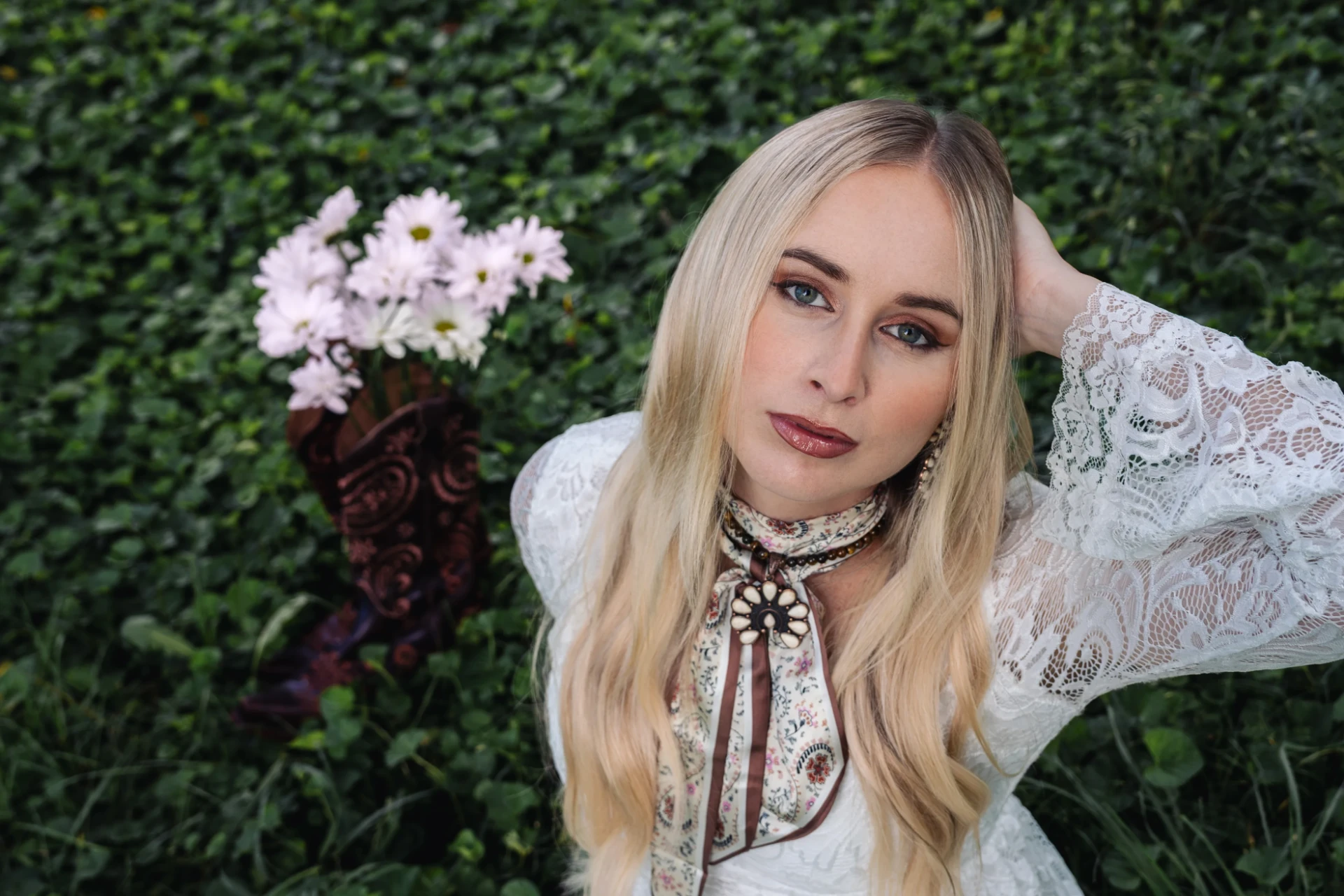 Melanie Dyer in white lace, lying in green clover beside a boot full of daisies